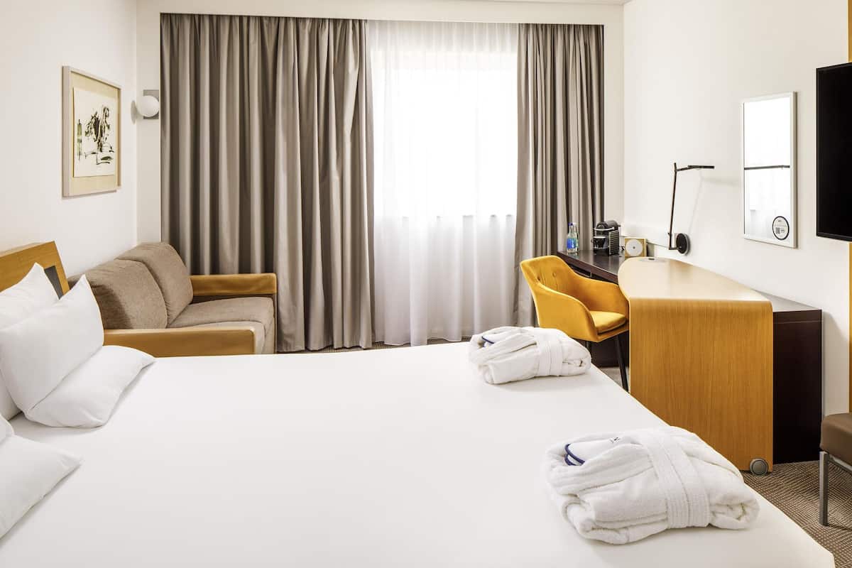 A hotel room with a bed, a desk, a chair, and a large window with curtains.