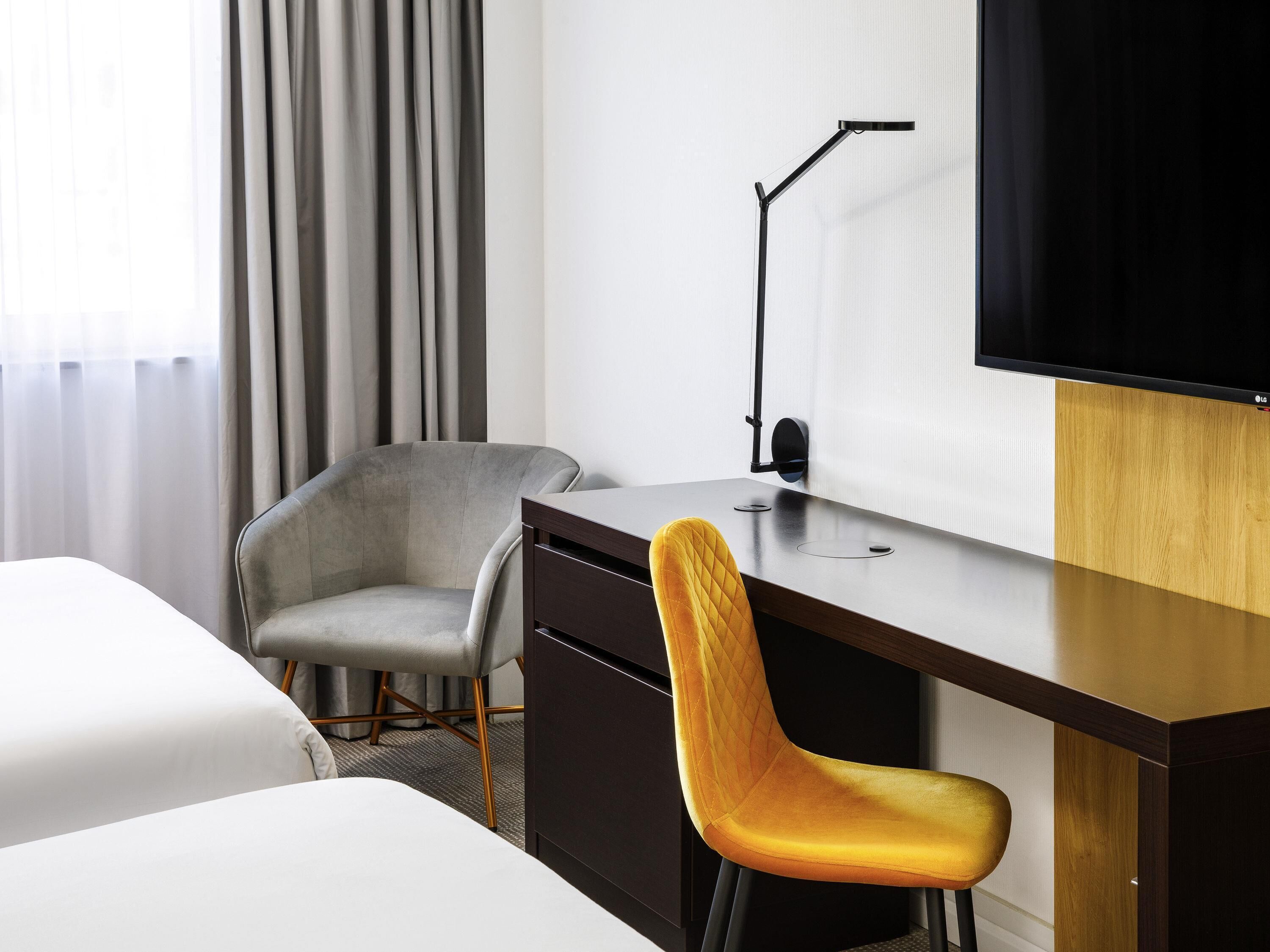A hotel room with a bed, a desk, a chair, a TV, and a window with curtains.