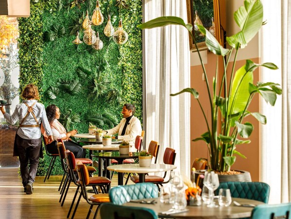 A restaurant with a green wall, hanging light fixtures, and a waiter serving a table. - Novotel London Greenwich (London)