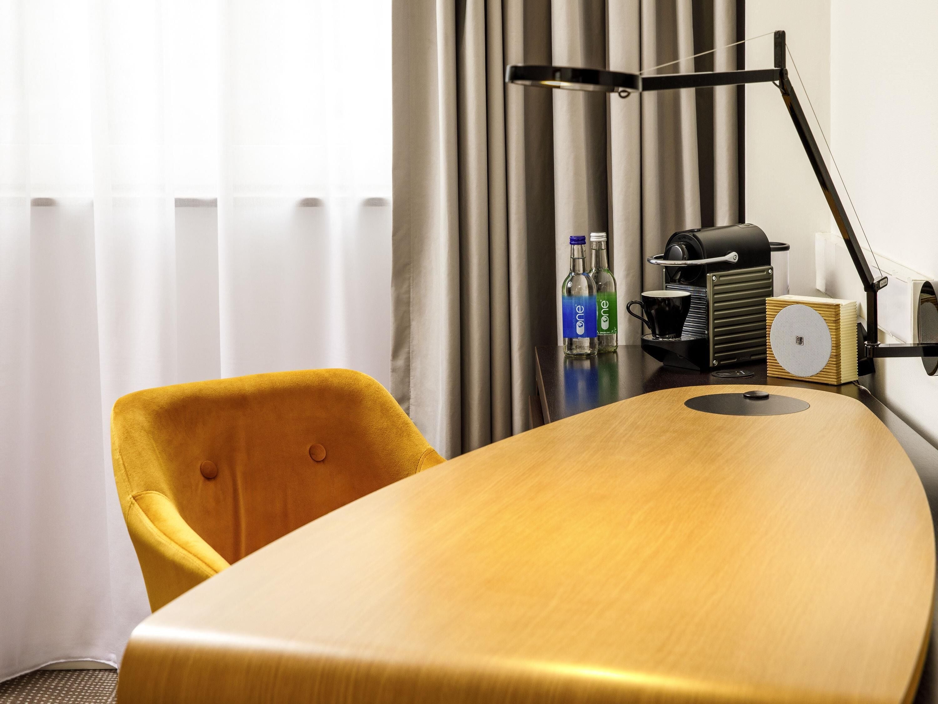 A hotel room with a wooden table, a yellow chair, a desk lamp, a coffee machine, and two bottles of water.