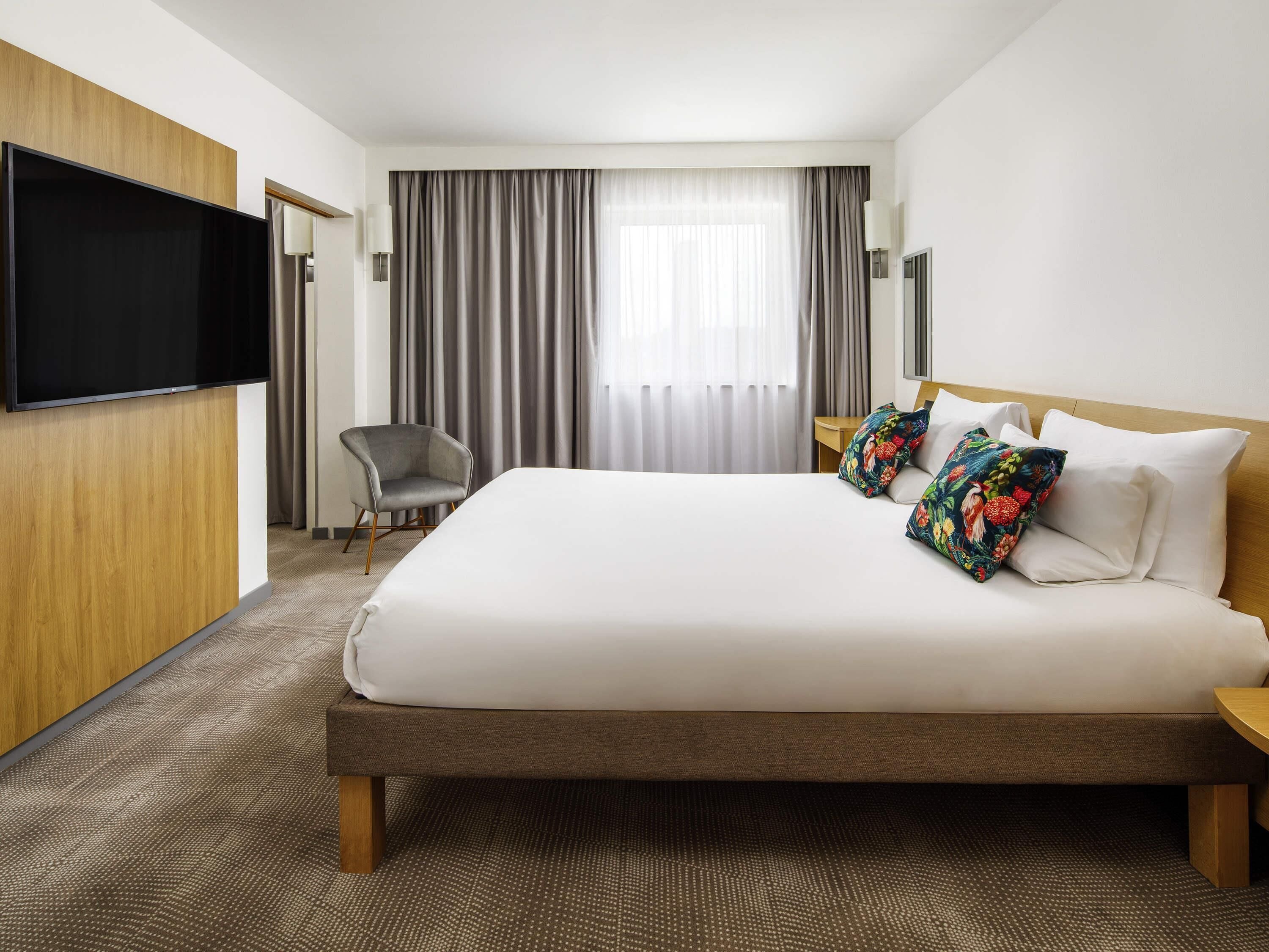 Een hotelkamer met een groot bed, een flatscreen televisie aan de muur, een stoel en een raam met gordijnen.