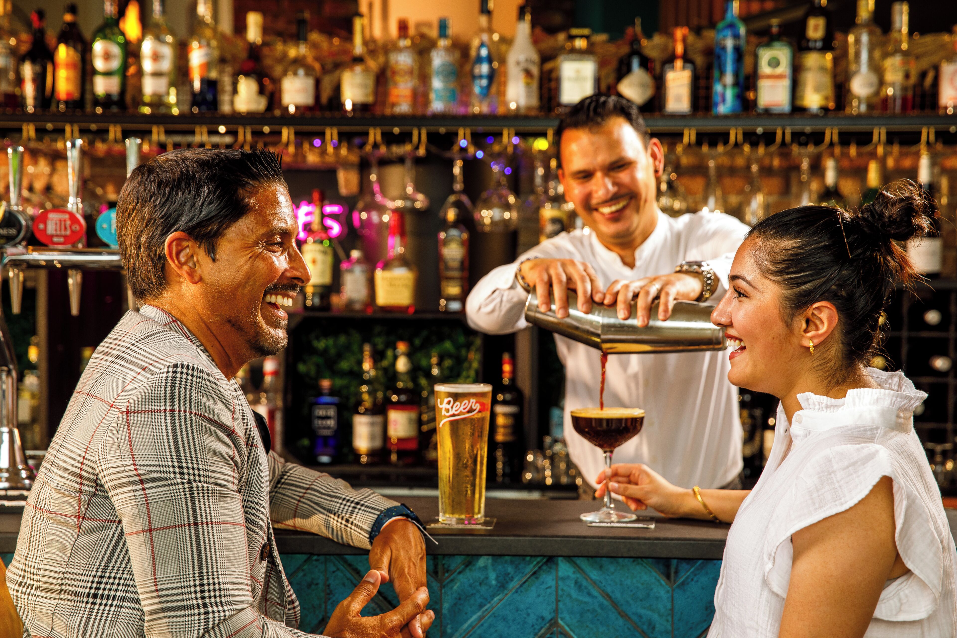 A bar with a bartender preparing drinks, a customer holding a beer, and another person in the background.