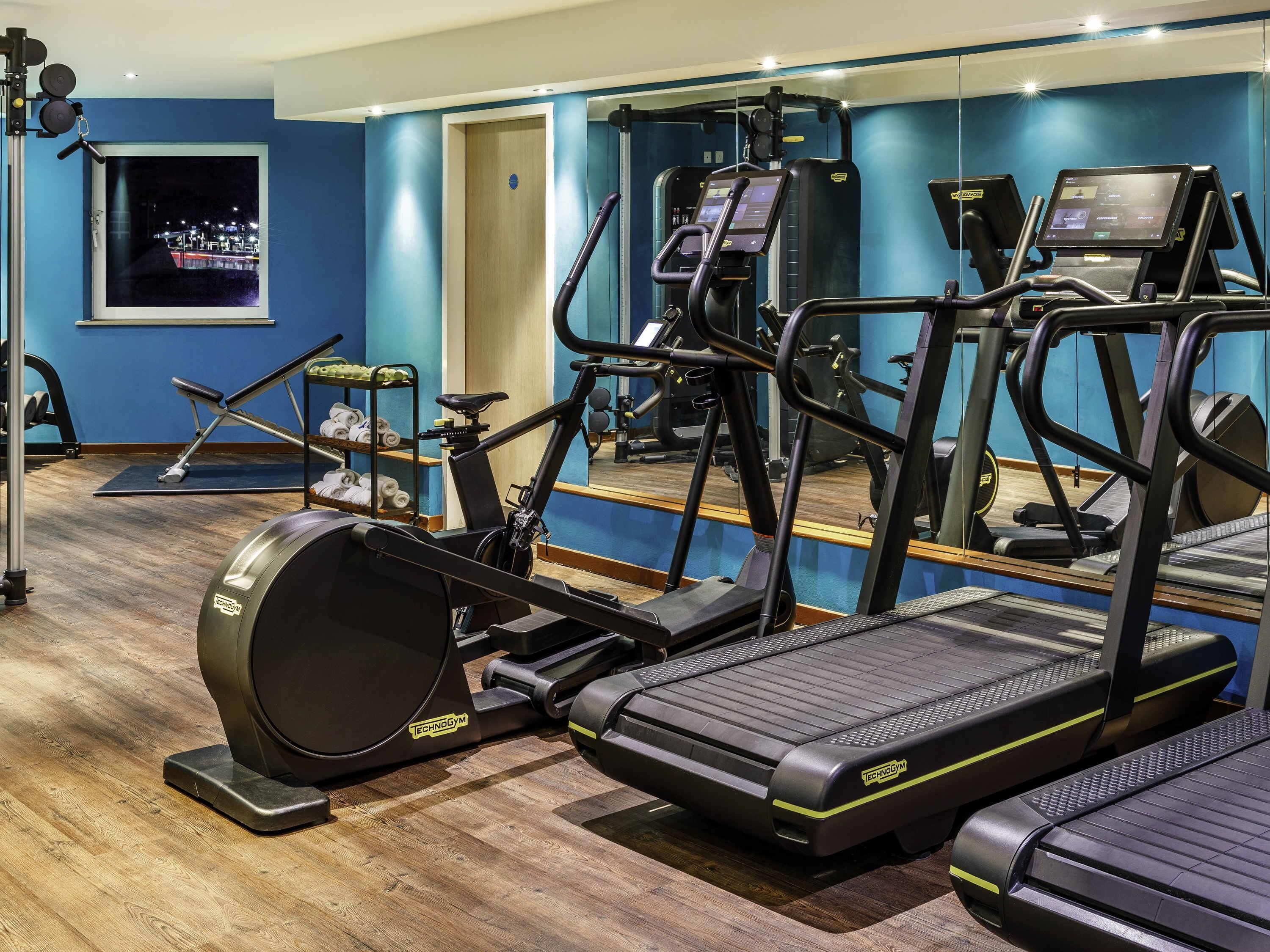 A gym with cardio equipment, a mirror, and a window with a city view.
