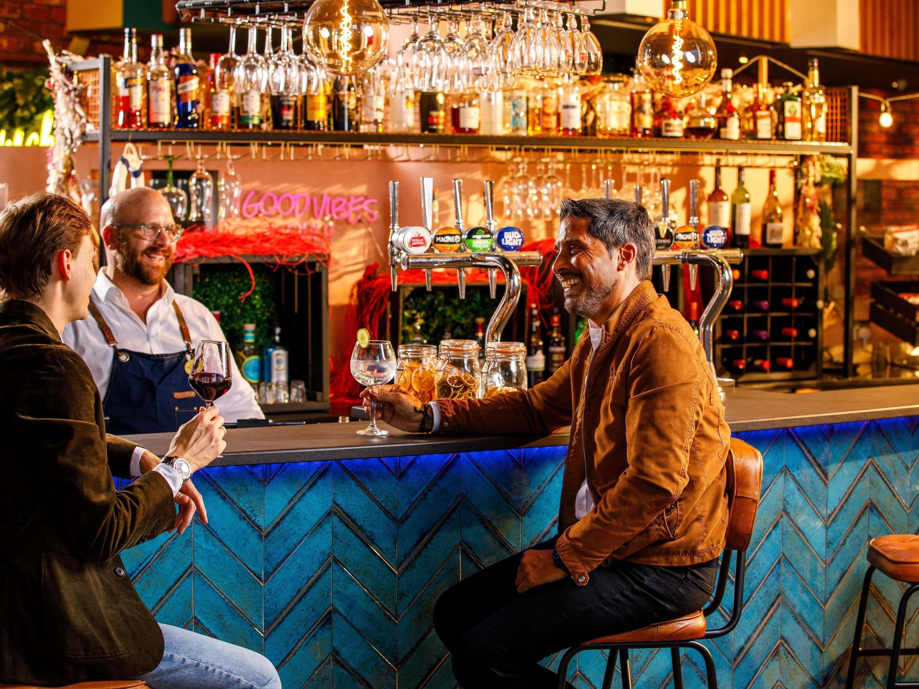 Um bar com parede em padrão azul Chevron, variedade de garrafas de bebidas e um letreiro com a inscrição 'GOOD VIBES'.