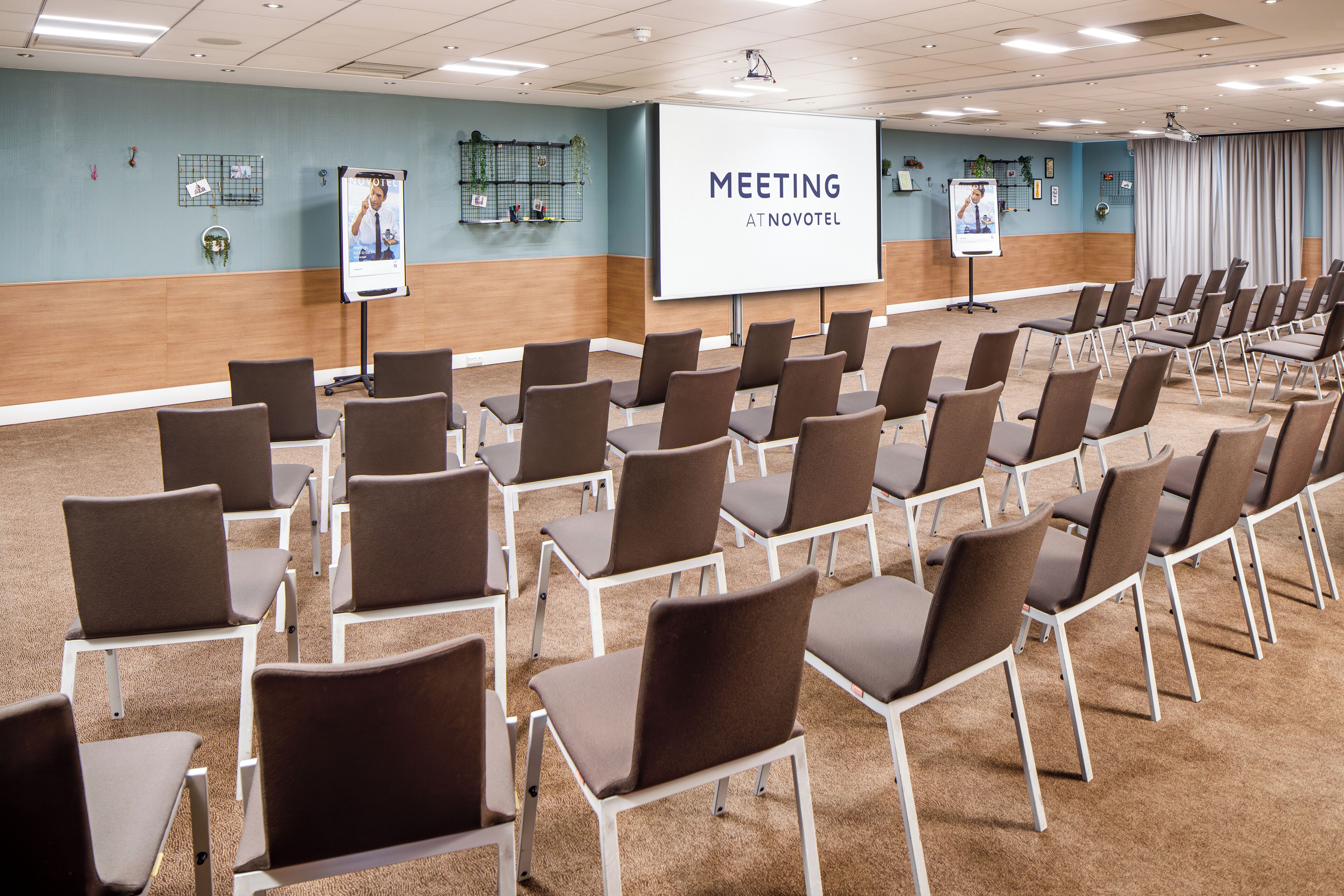 Una sala riunioni con file di sedie, un grande schermo e uno striscione con su scritto 'MEETING AT NOVOTEL'.