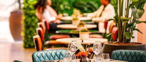 Ein Restaurant mit runden Tischen, grünen Sitzstühlen und einer üppigen Grünkulisse.