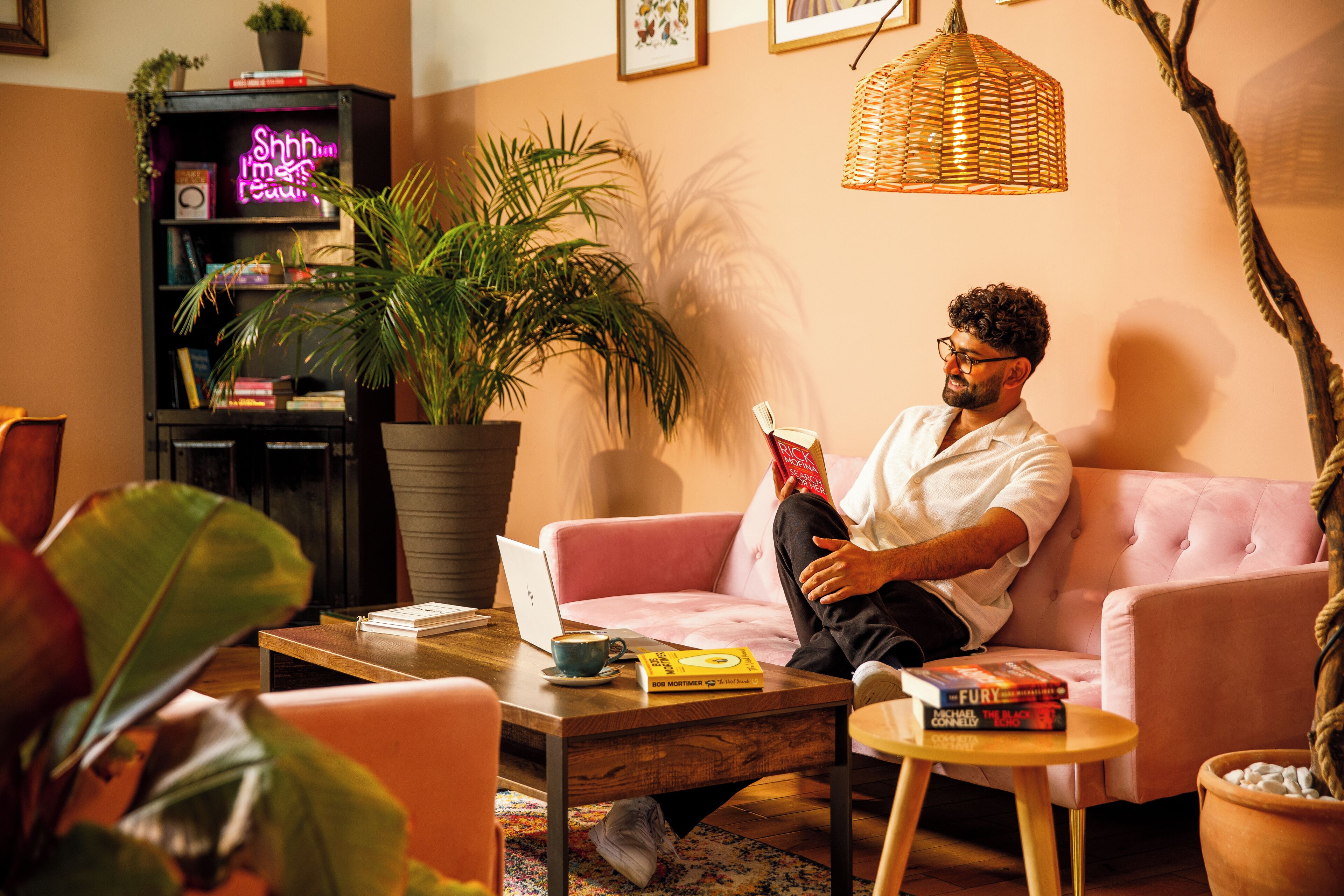 Ein gemütliches Wohnzimmer mit einem rosa Sofa, einem Couchtisch mit Büchern und einem Laptop, einem Bücherregal mit verschiedenen Gegenständen und einer großen Topfpflanze.