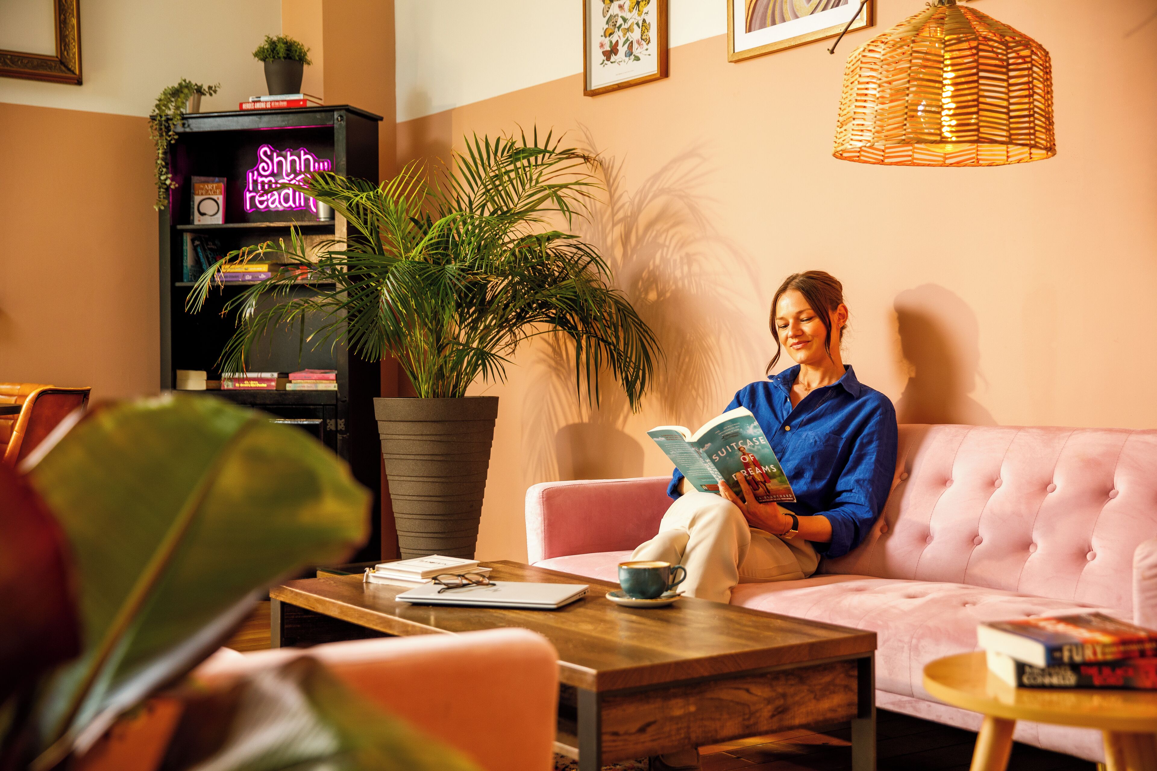 A woman reading a book in a cozy living room with a pink sofa, a wooden coffee table, and a bookshelf with a neon sign.