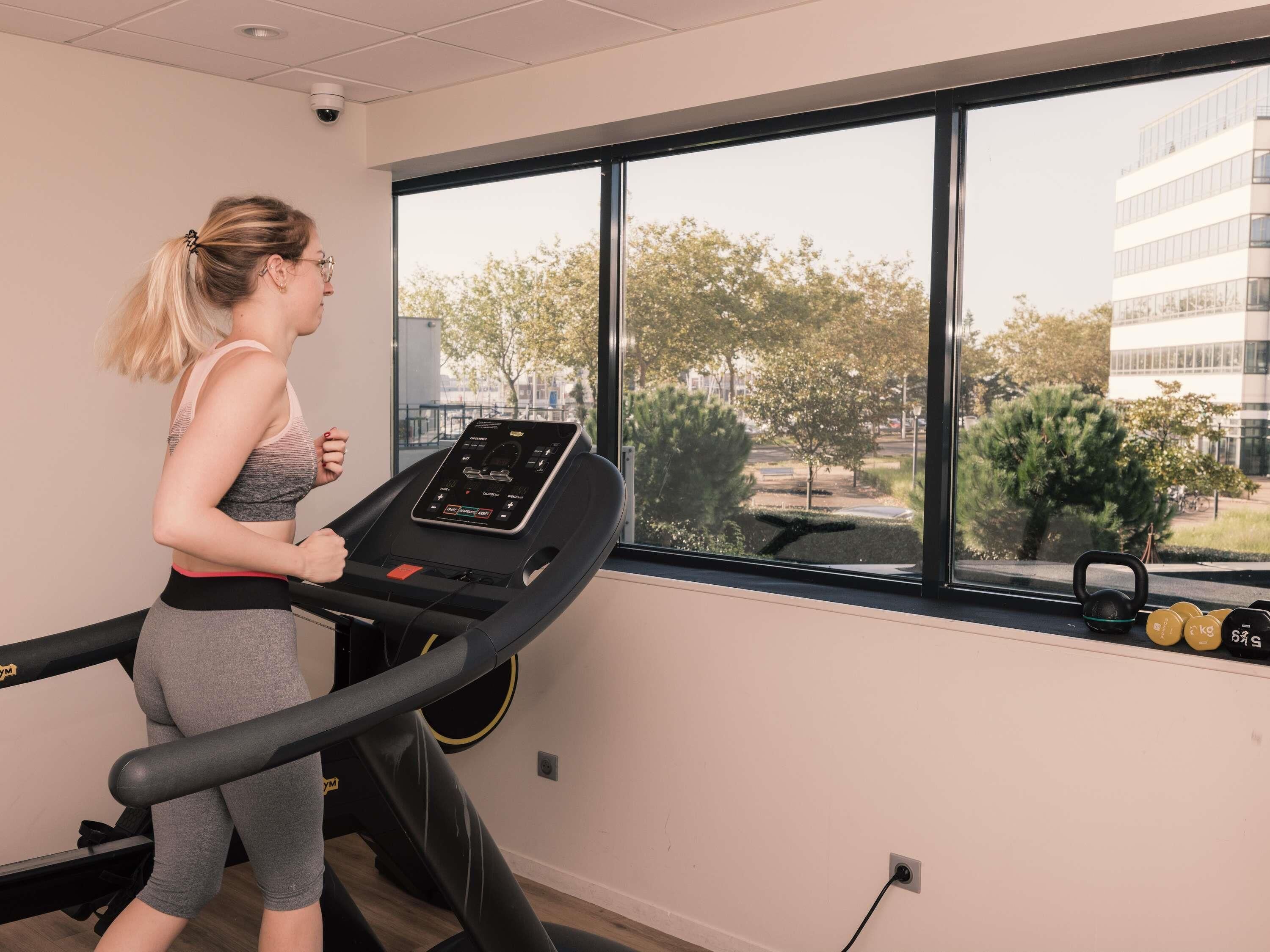 Une femme court sur un tapis de course dans une salle de sport, avec une fenêtre donnant sur des arbres et des immeubles.