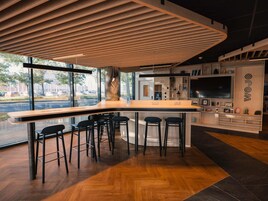 Uma área de bar moderna com teto de ripas de madeira, banquetas, uma televisão grande e vista para a rua.