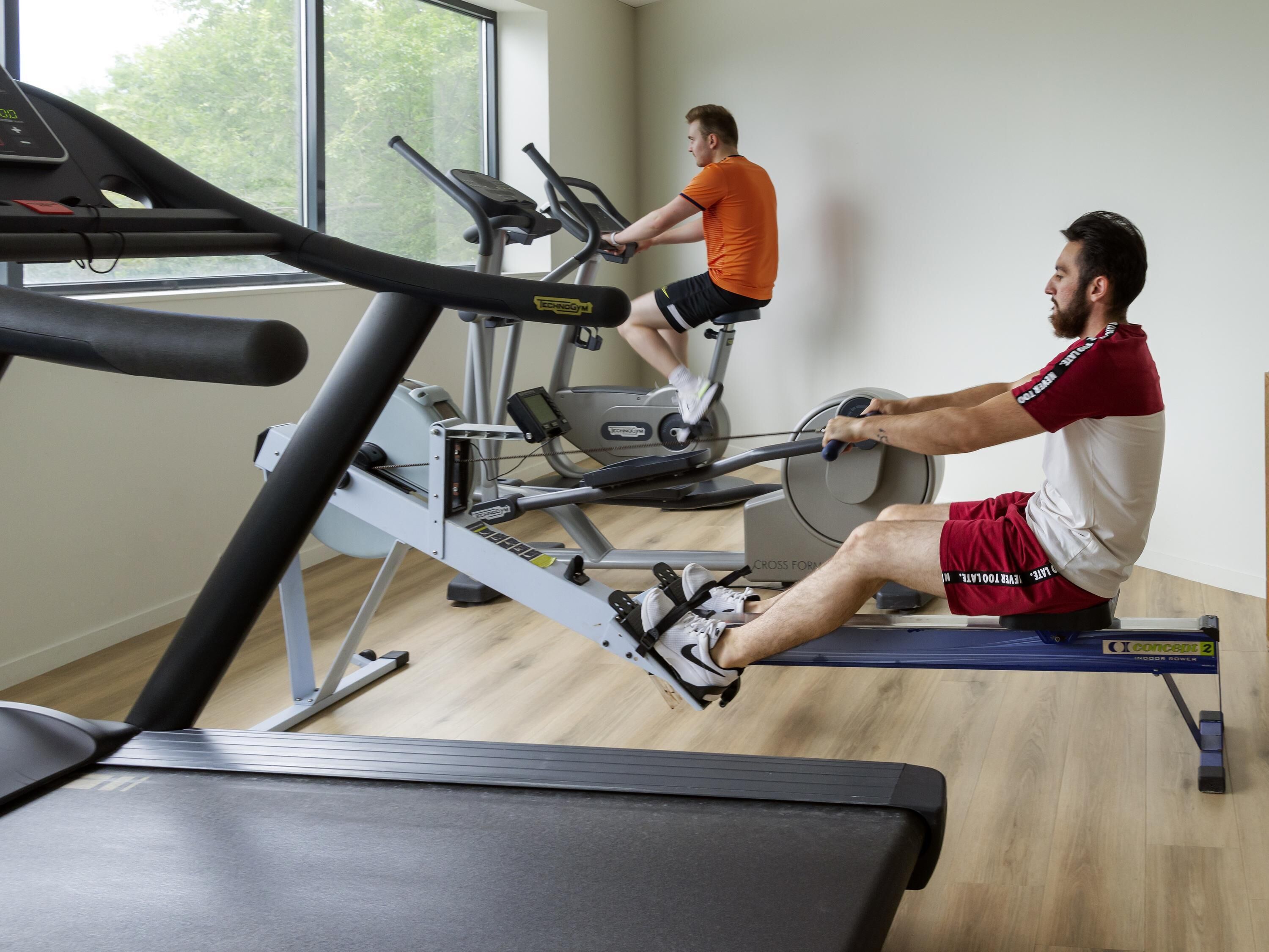 A gym with stationary bikes and treadmills, wooden flooring, and large windows.