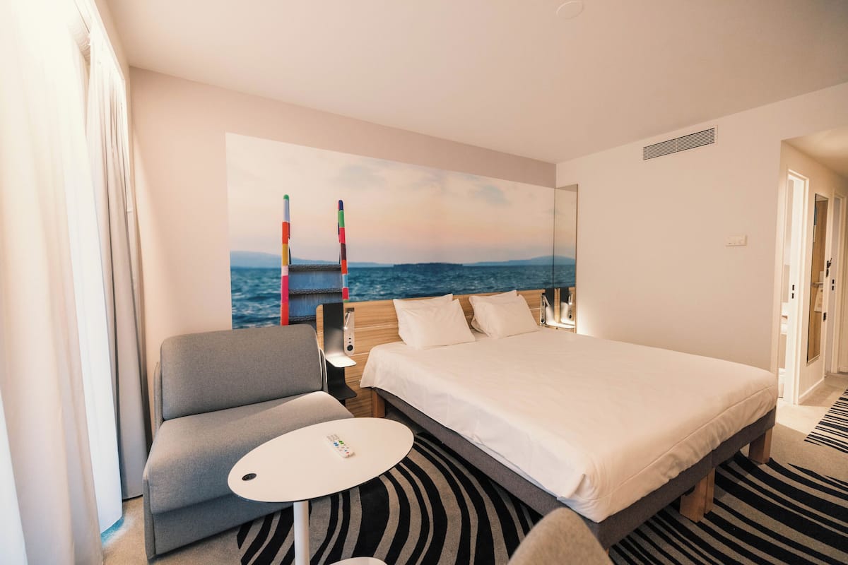 A hotel room with a bed, a gray armchair, a small round table, and a large painting of a coastal scene.