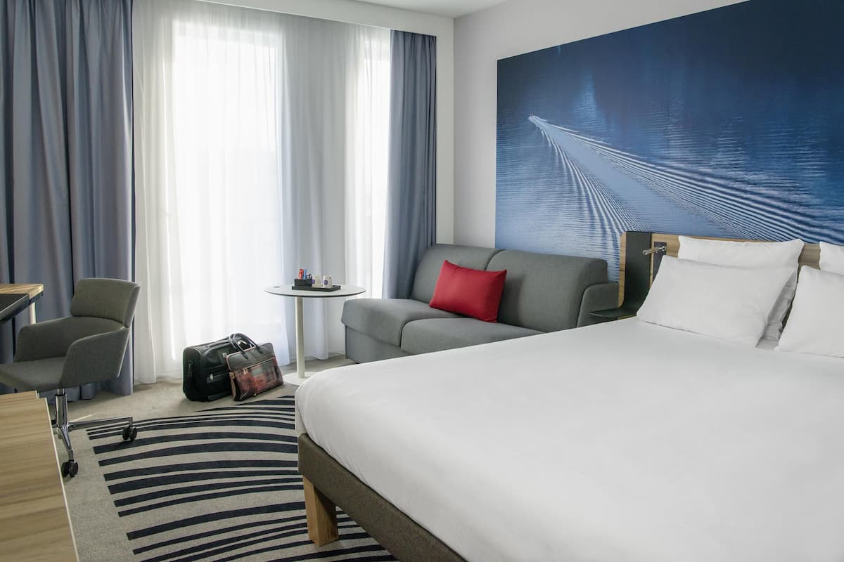 A hotel room with a bed, a grey sofa with a red pillow, a grey armchair, a small round table with a lamp, and a large window with curtains.