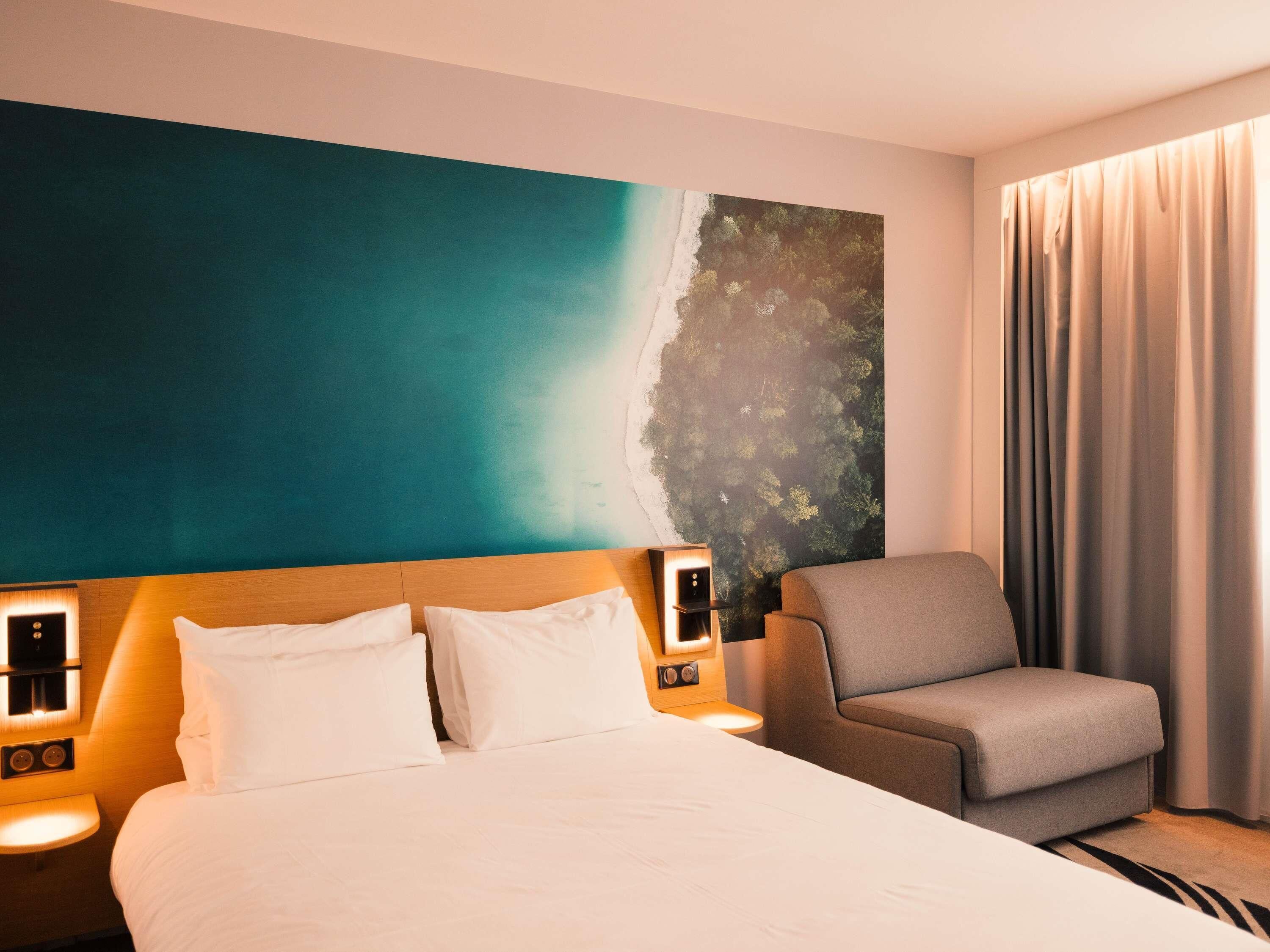 A hotel room with a bed, a chair, a nightstand, and a large wall mural.