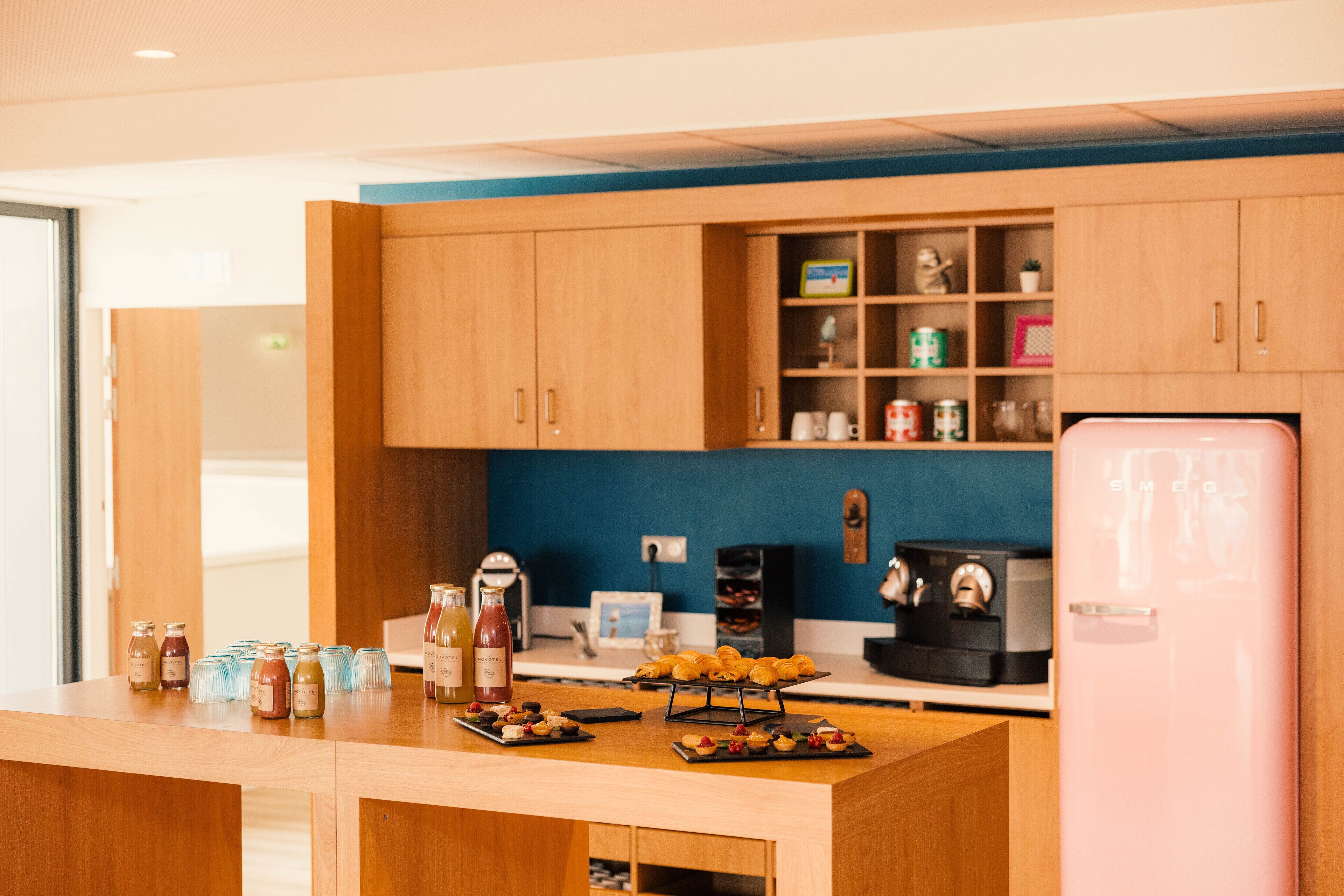 A modern kitchen with wooden cabinets, a countertop with food items, a coffee machine, and a pink refrigerator.