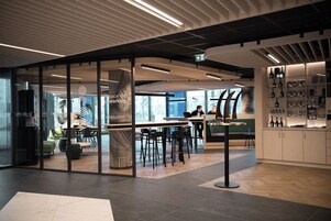 Um espaço de coworking moderno, com divisórias de vidro, piso de madeira e diversas opções de assentos.
