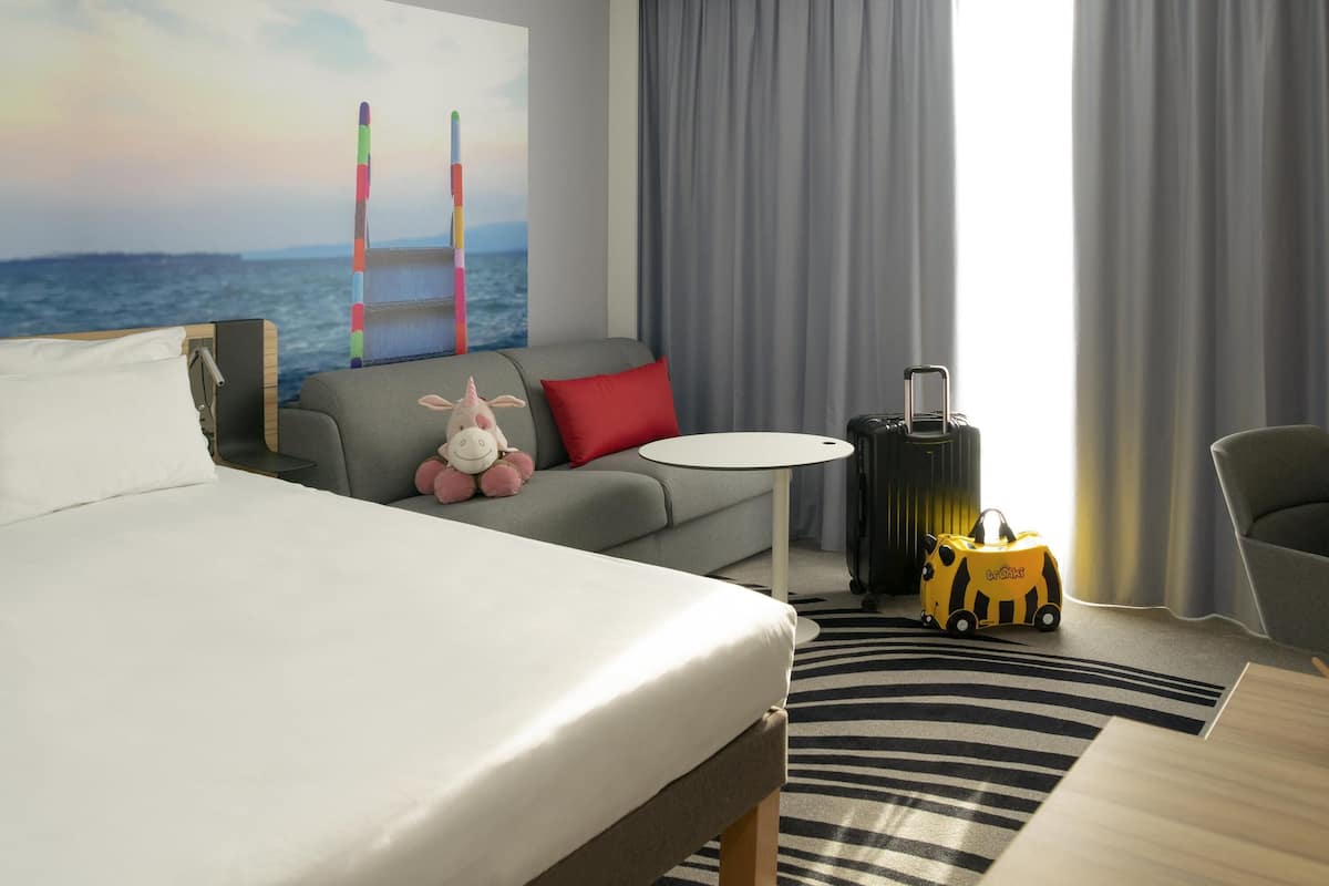 A hotel room with a bed, a sofa, a small table, a suitcase, a toy, and a view of the sea through curtains.