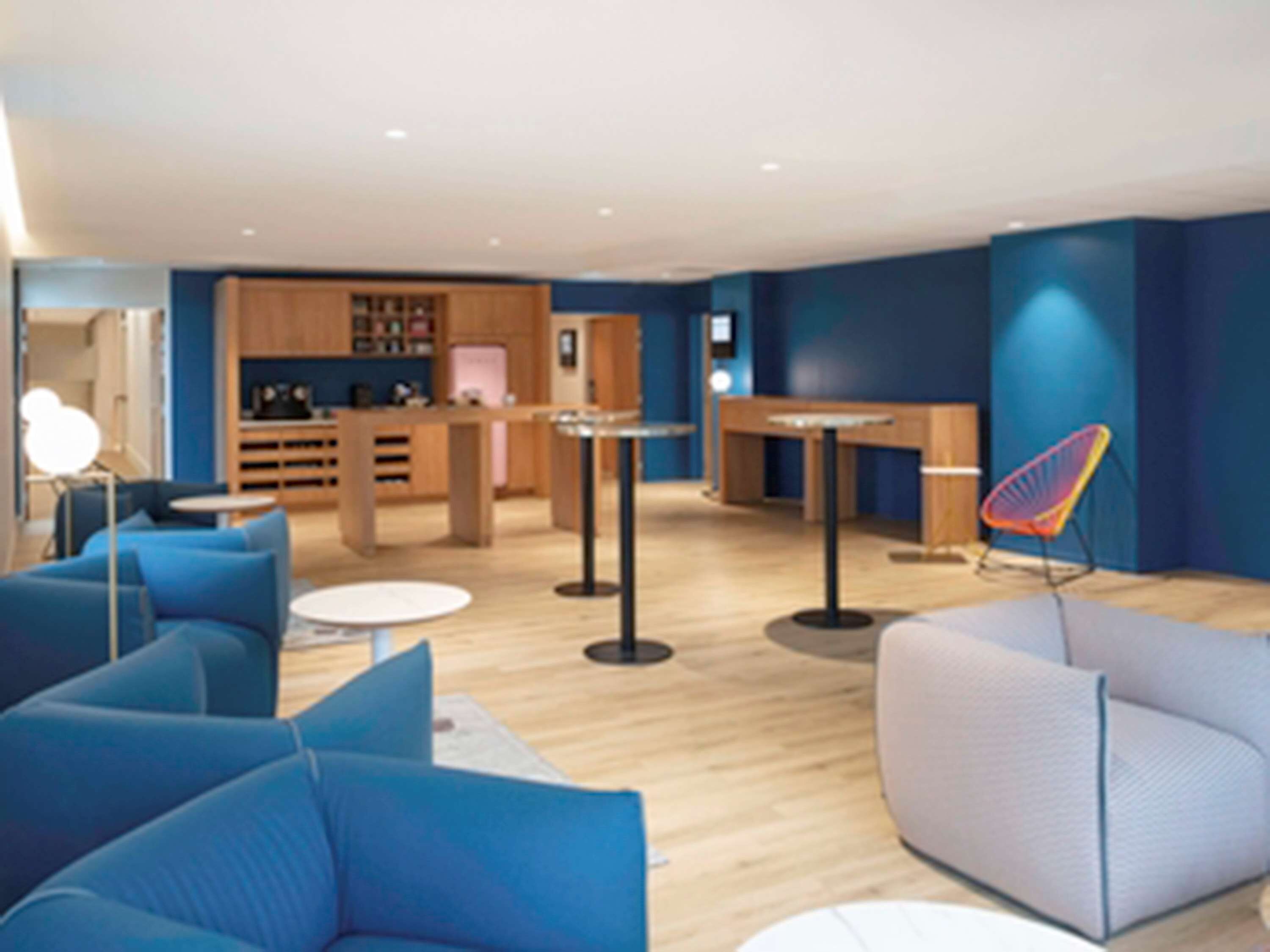 A modern interior with blue and grey sofas, a small kitchen area, and a dining table.