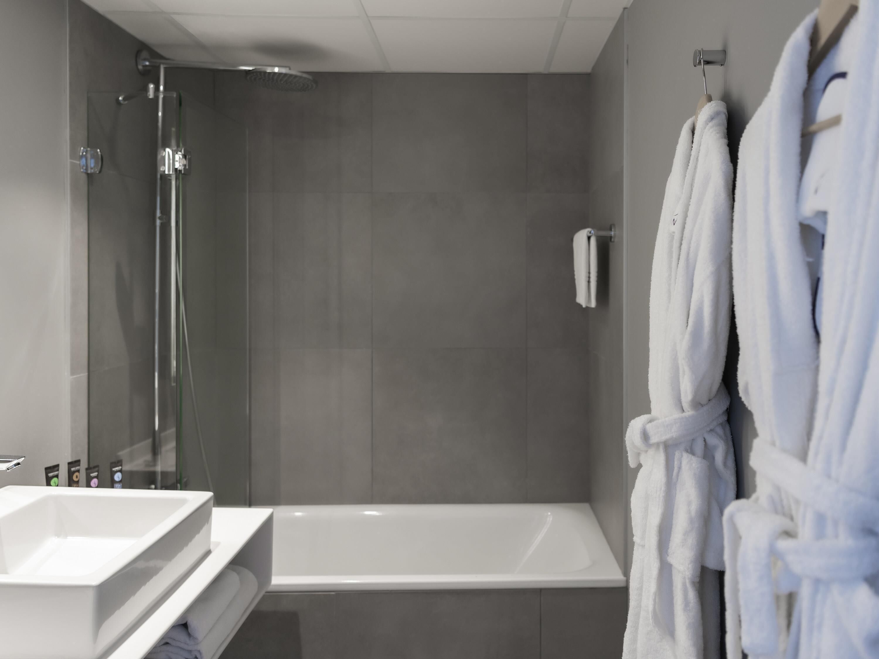Een moderne badkamer met een douchecabine van glas, een wit bad en twee witte badjassen aan haken.