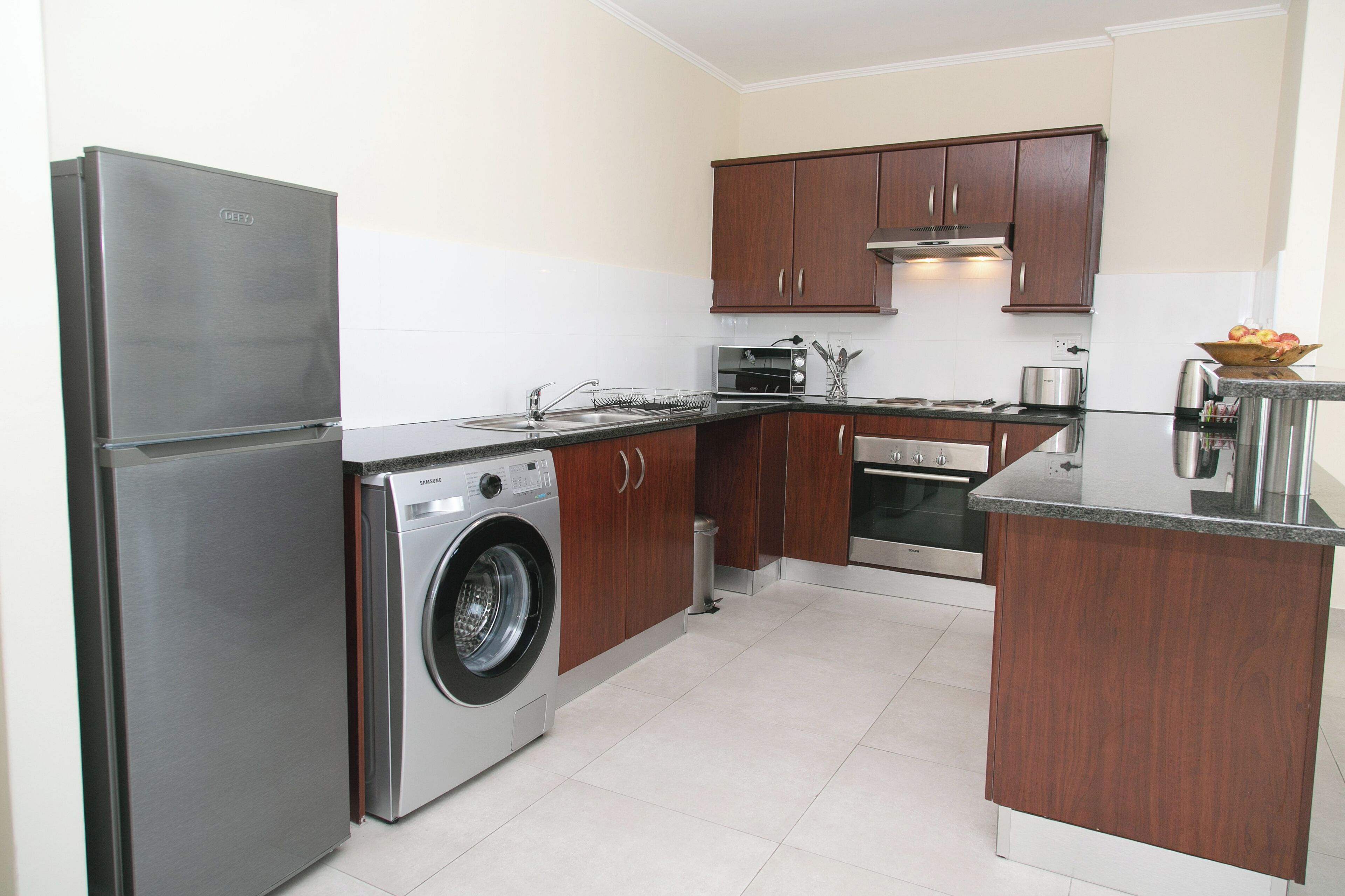 A compact kitchen with a stainless steel refrigerator, a washing machine, wooden cabinets, and a small countertop.