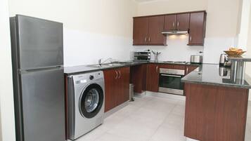 A compact kitchen with a stainless steel refrigerator, a washing machine, wooden cabinets, and a small countertop.