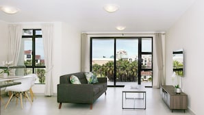 A modern living room with a sofa, dining table, and chairs, a TV, and a balcony with a view of buildings.