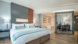 A modern hotel room with a large bed, bedside tables, a bathroom with a shower, and a dining area with a table and chairs.