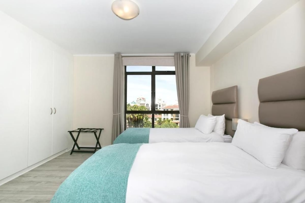 A hotel room with two beds, a window with curtains, a small table, and a view of buildings outside.