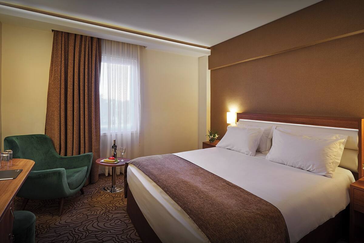 Een hotelkamer met een groot bed, een bureau met stoel, een klein tafeltje met fles en glas, en een raam met gordijnen.