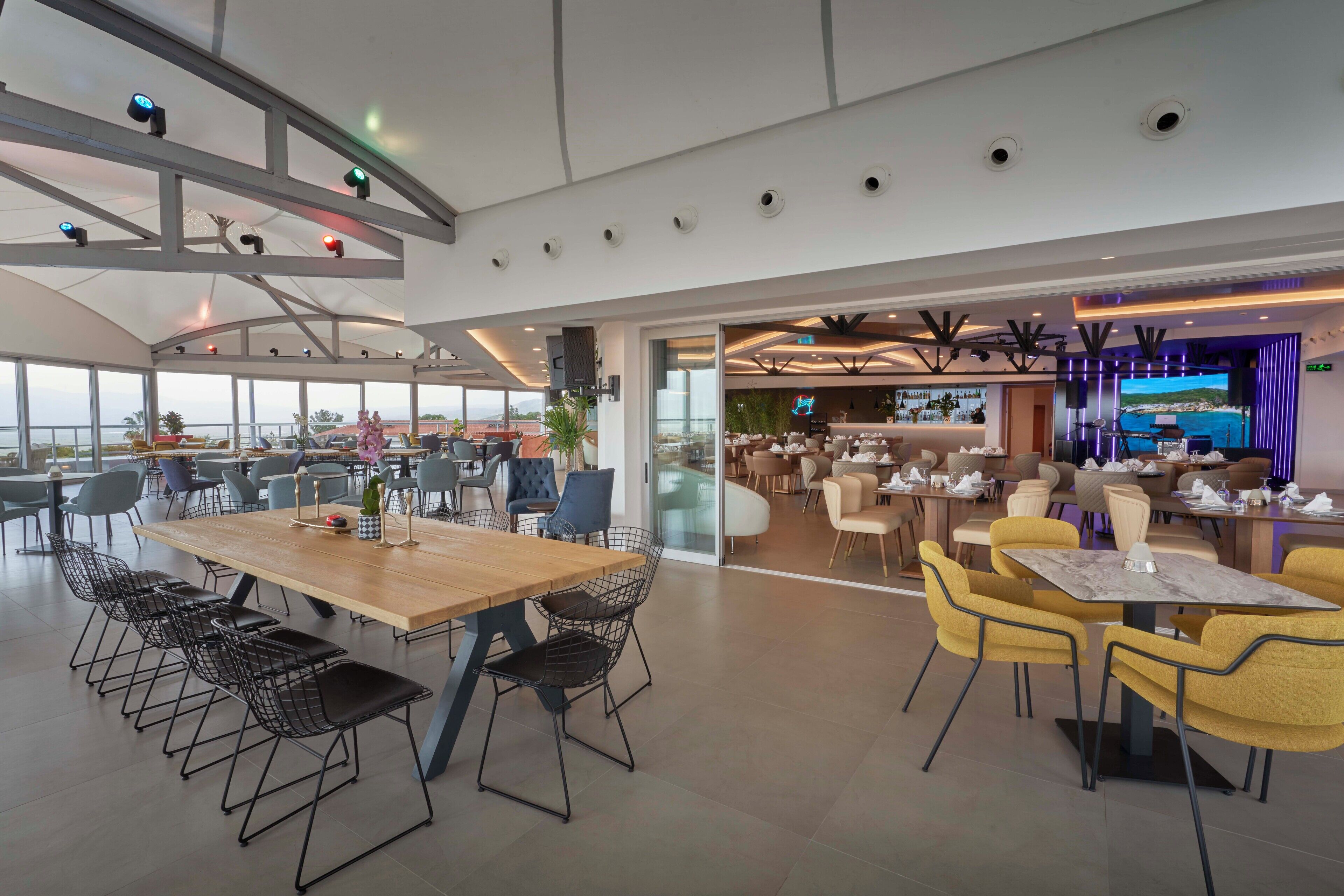 Un comedor amplio con varias mesas y sillas, una zona de bar con una pantalla grande, y vistas al exterior.