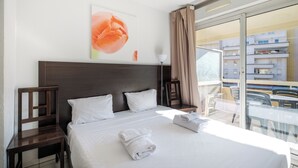 A hotel room with a large bed, two pillows, a wooden headboard, a white wall with a flower painting, a glass balcony with chairs, and a view of buildings.