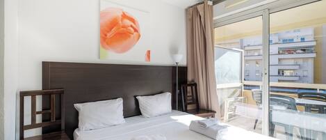 Habitación de hotel con una cama grande, dos almohadas, cabecero de madera, pared blanca con una pintura floral, balcón de vidrio con sillas y vistas a edificios.