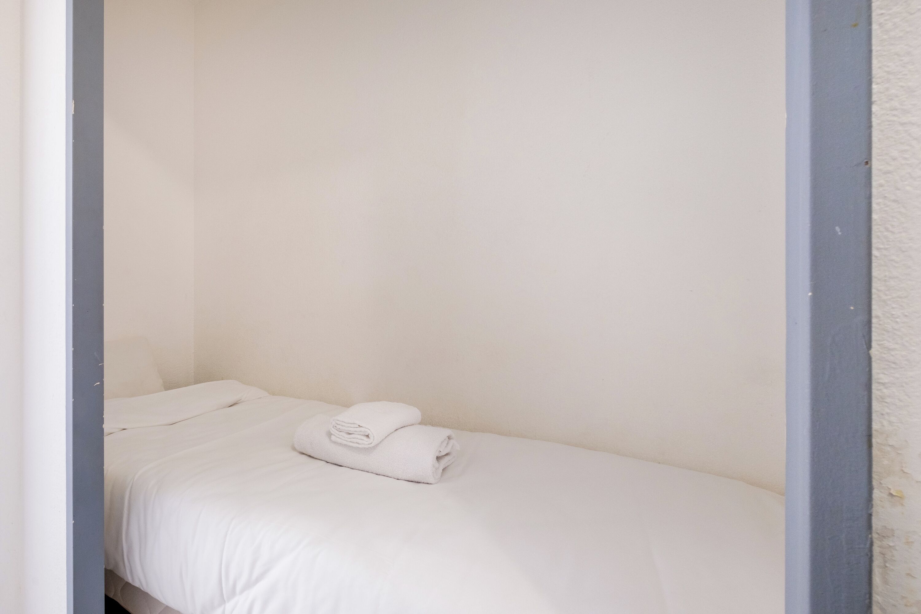 Ein kleines Zimmer mit einem Einzelbett, weißer Bettwäsche und einem ordentlich gefalteten Handtuch auf dem Bett.