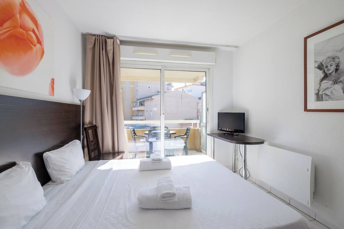 Hotellihuone, jossa on sänky, televisio, pieni pöytä, parveke tuoleineen ja näkymä rakennuksiin.