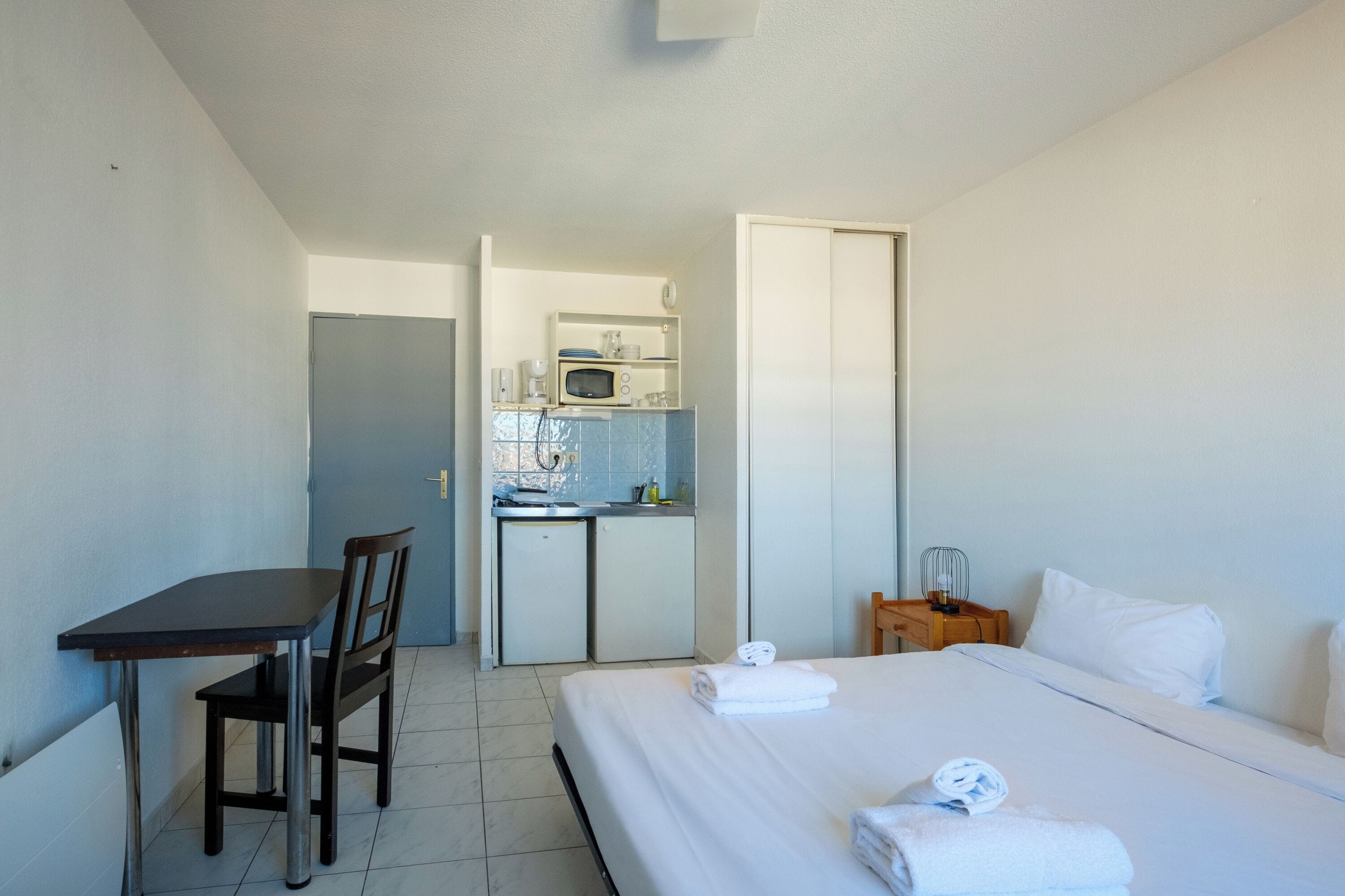 A hotel room with a bed, a desk, a chair, a kitchenette, and a bedside table.