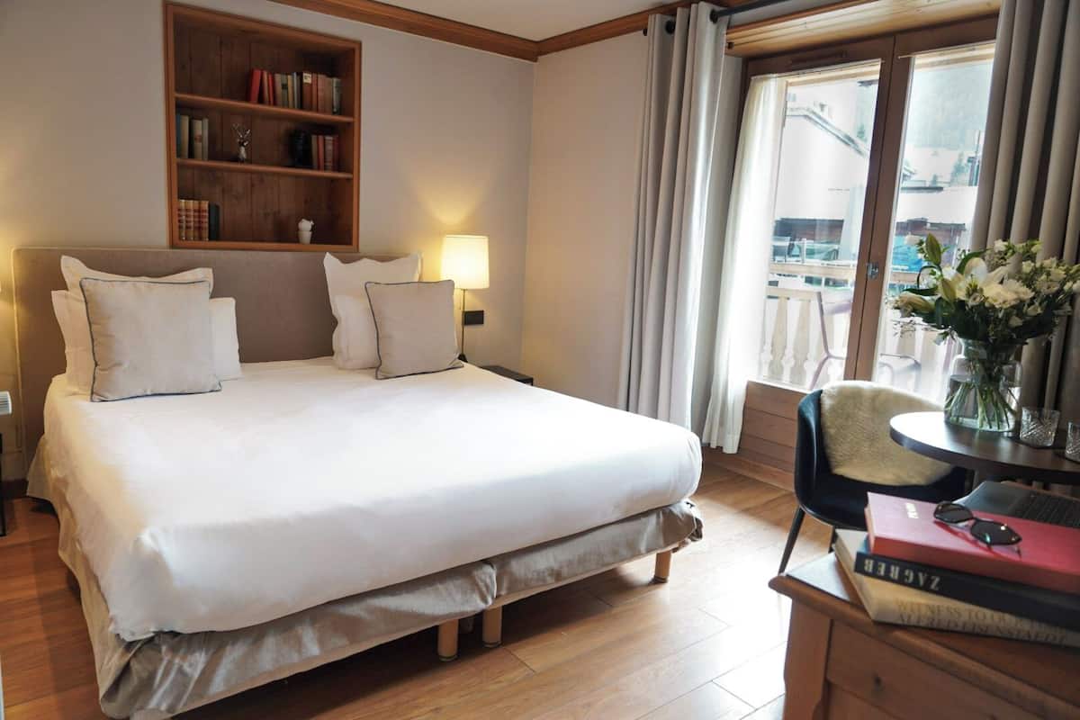Habitación de hotel con una cama grande, una mesita de noche con lámpara, una estantería y vistas al exterior a través de una ventana.
