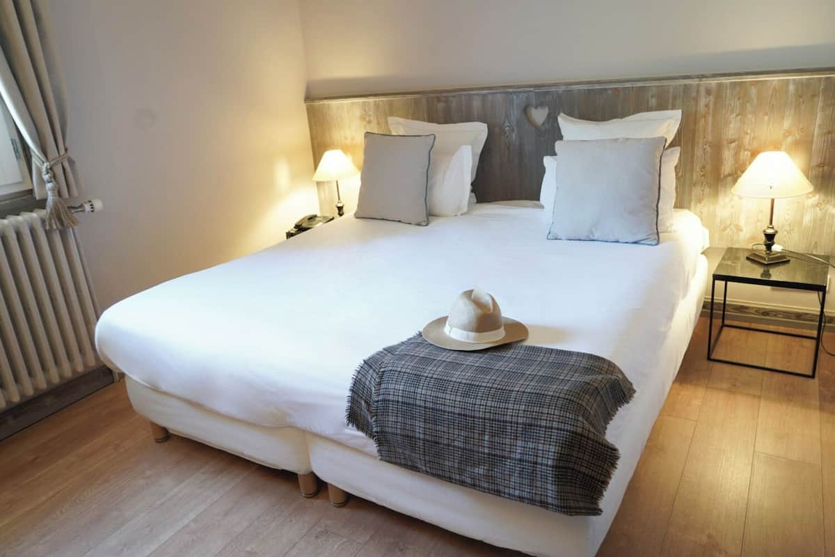 Una cama bien hecha con sábanas blancas, una manta de cuadros y un sombrero sobre la cama. Hay dos lámparas de noche, un cabecero de madera y un radiador a la izquierda.