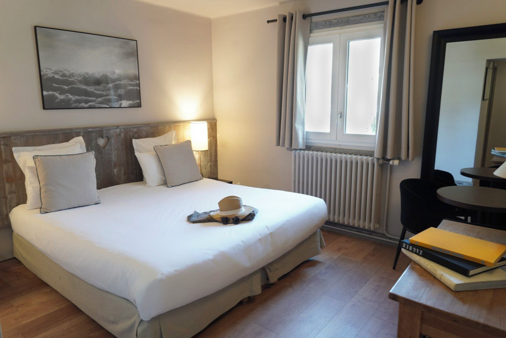 A hotel room with a large bed, a bedside table with books, a window with curtains, a radiator, and a wall-mounted picture.