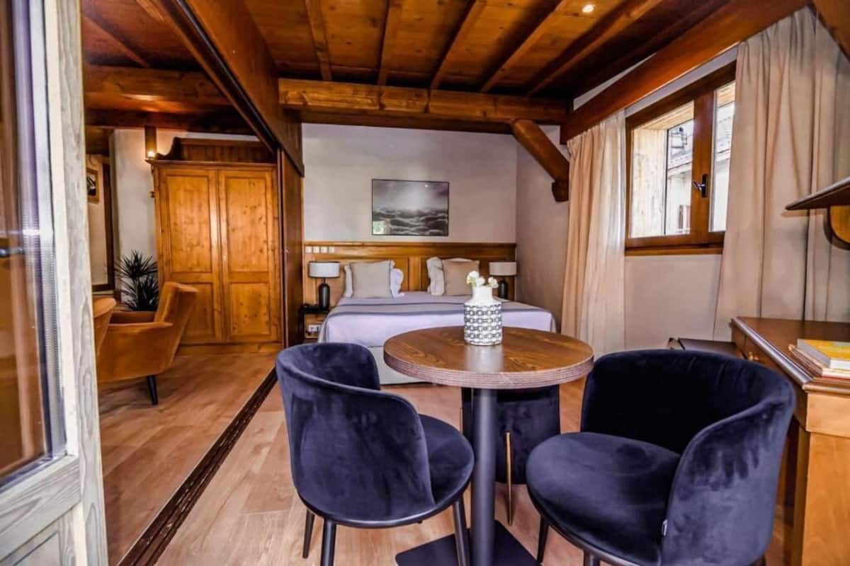 Una habitación acogedora con techo de madera, una cama con sábanas blancas, dos sillas negras alrededor de una mesa redonda pequeña y un ventanal con cortinas.