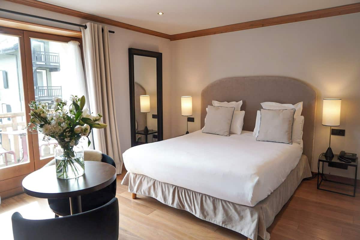 Habitación de hotel con una cama grande, dos lámparas de noche, una mesa redonda con una silla, un florero con flores y vistas al balcón exterior.