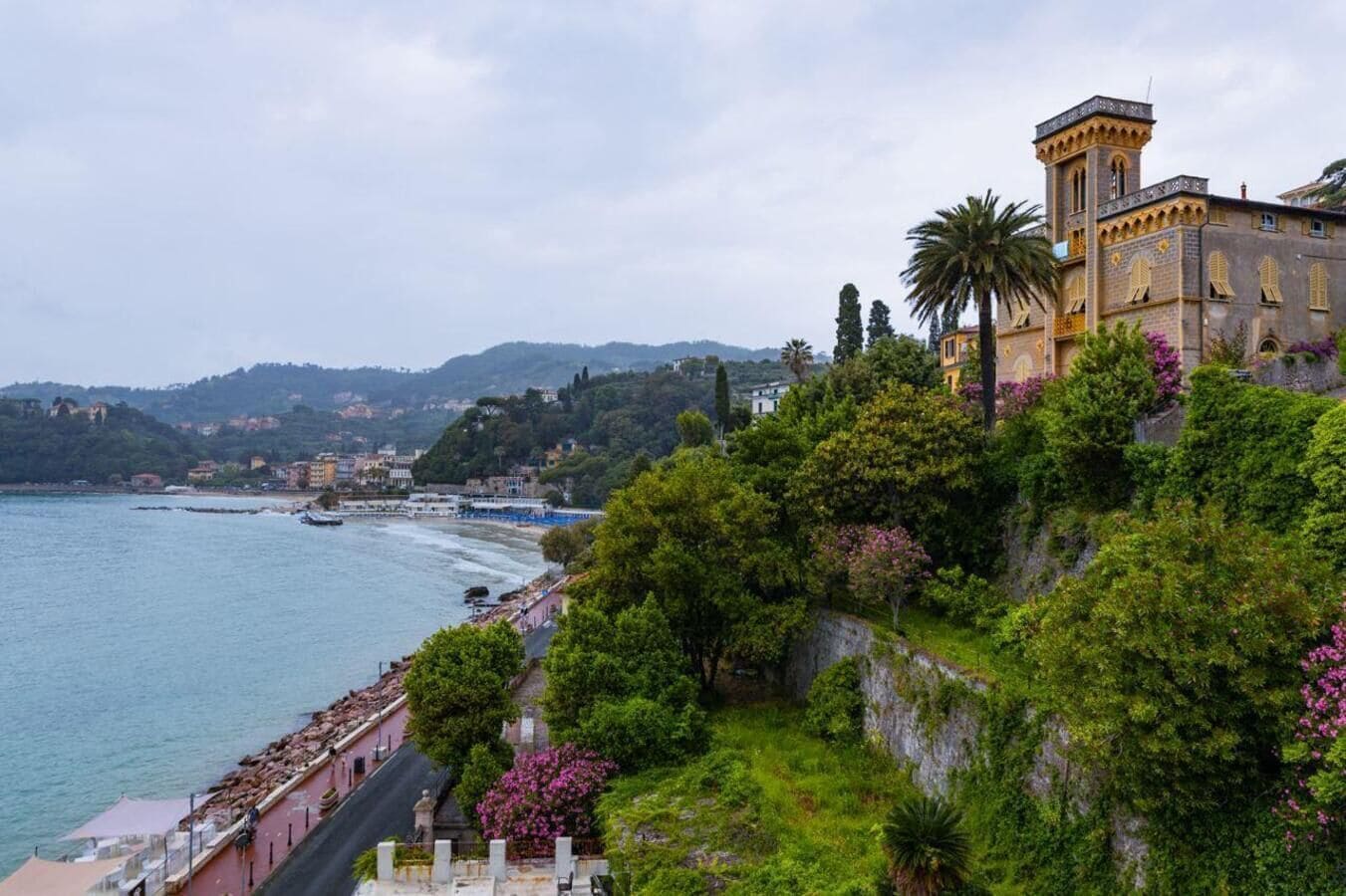 A coastal building with a tower, surrounded by lush greenery and overlooking a beach.