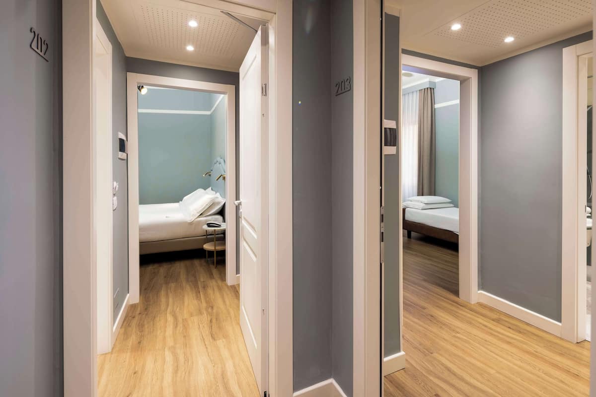 Un corridoio d'hotel con pavimento in legno, un letto con materasso bianco e un comodino, e uno specchio a parete.