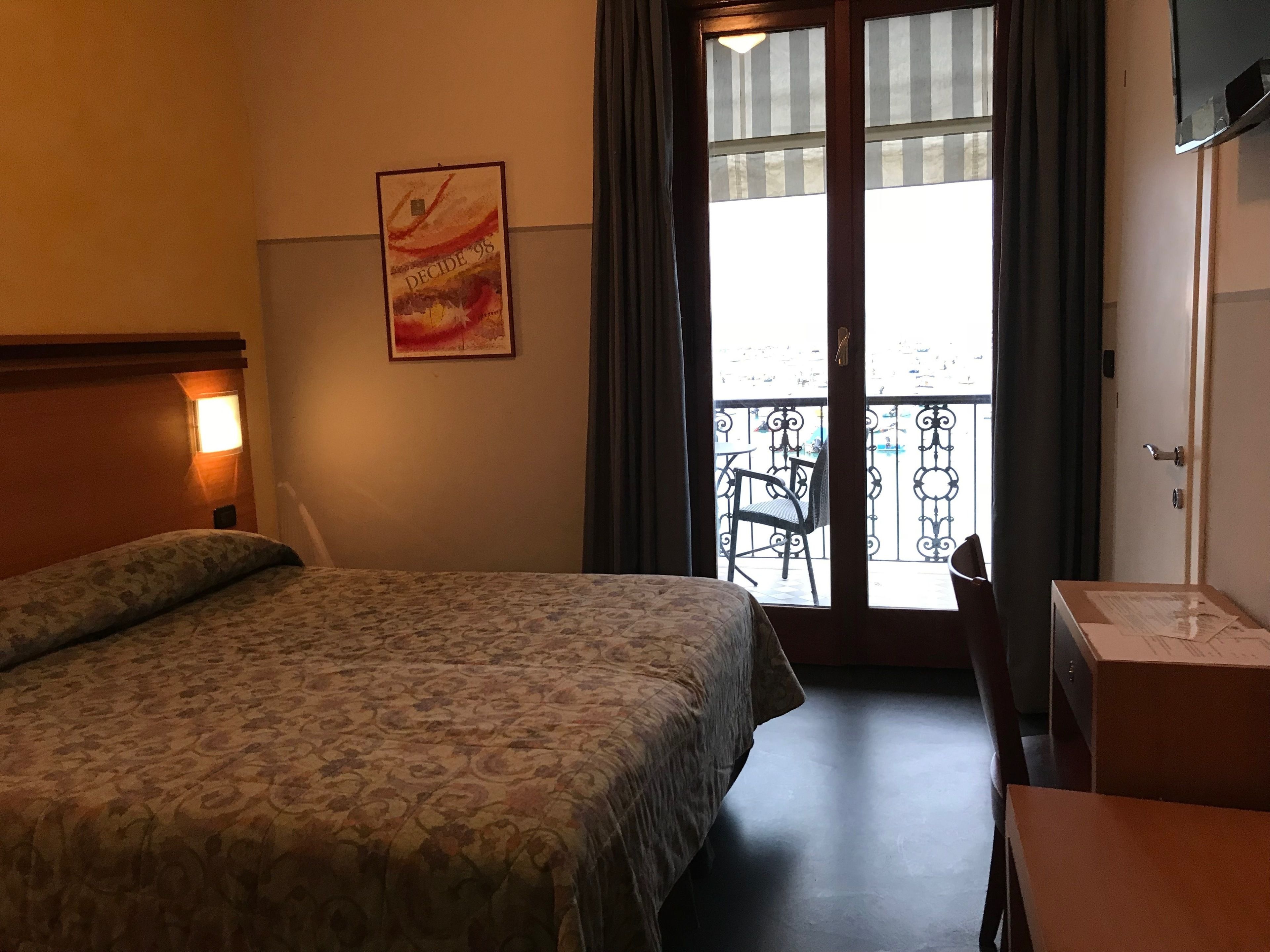 A hotel room with a bed, a desk, a chair, a balcony with a view, and a poster on the wall.