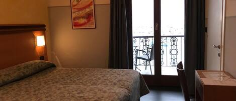 A hotel room with a bed, a desk, a chair, a balcony with a view, and a poster on the wall.