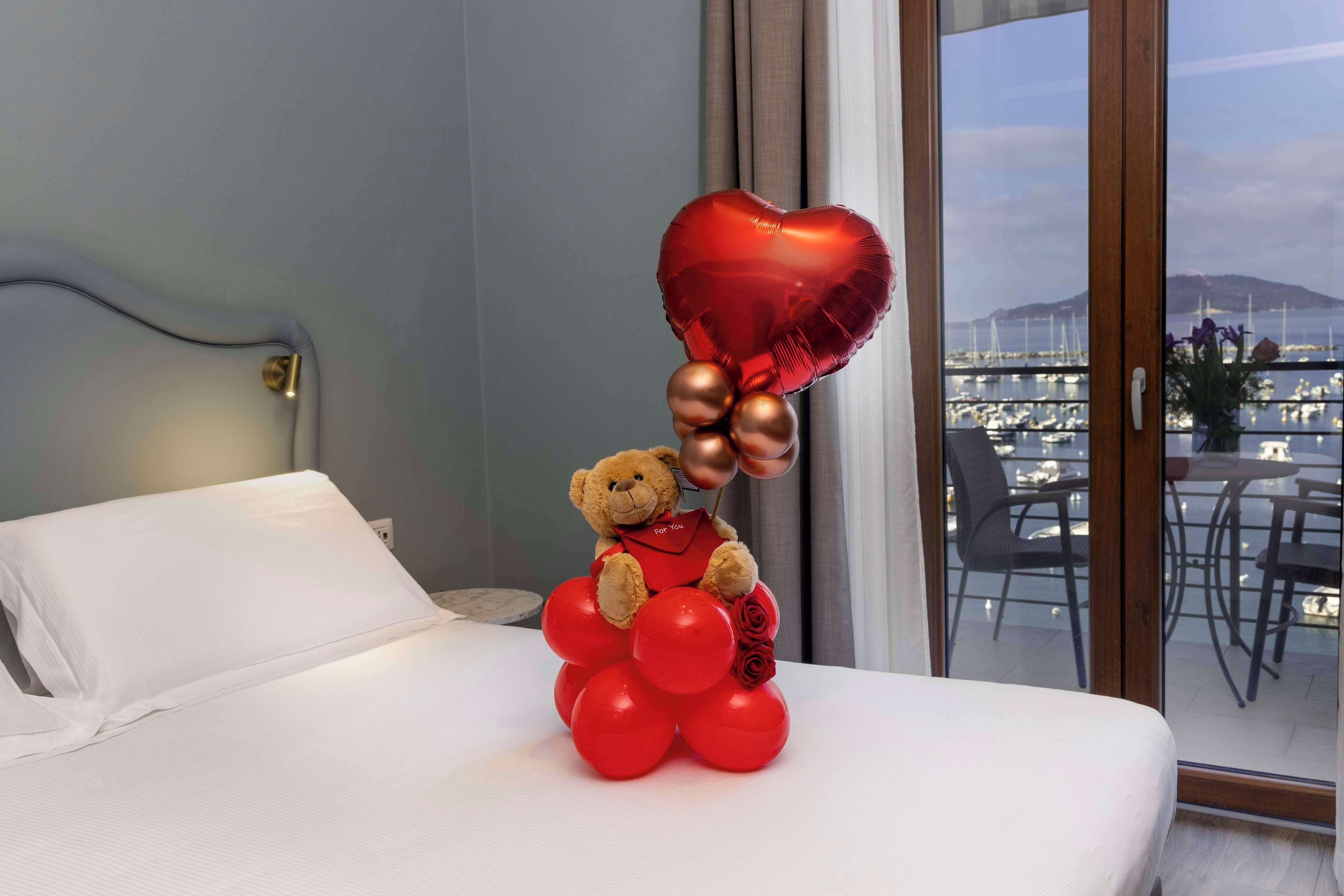 A bed with a teddy bear holding a heart-shaped balloon and red balloons, a window with a view of a marina, and a white pillow.