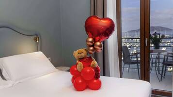 Ein Bett mit einem Teddybären, der einen herzförmigen Luftballon und rote Ballons hält, ein Fenster mit Blick auf einen Hafen und ein weißes Kopfkissen.