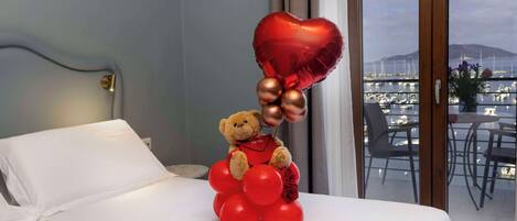 A bed with a teddy bear holding a heart-shaped balloon and red balloons, a window with a view of a marina, and a white pillow.