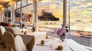 Een restaurant met gedekte tafels, grote ramen met uitzicht op een historisch gebouw en een jachthaven.