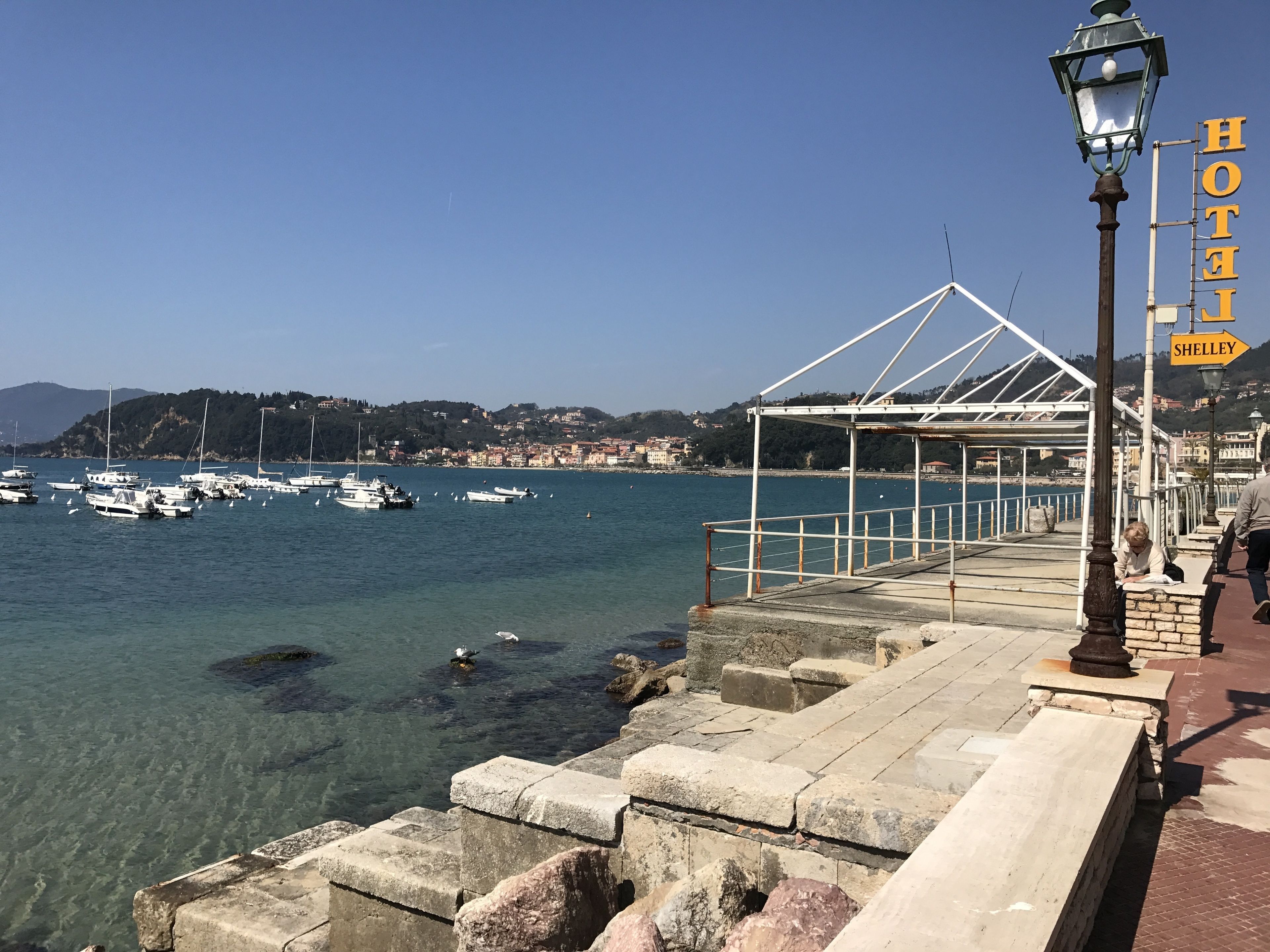 A waterfront promenade with a clear view of boats docked in the background, a stone pathway leading to a covered area, and a sign indicating the location as Hotel Shelley.