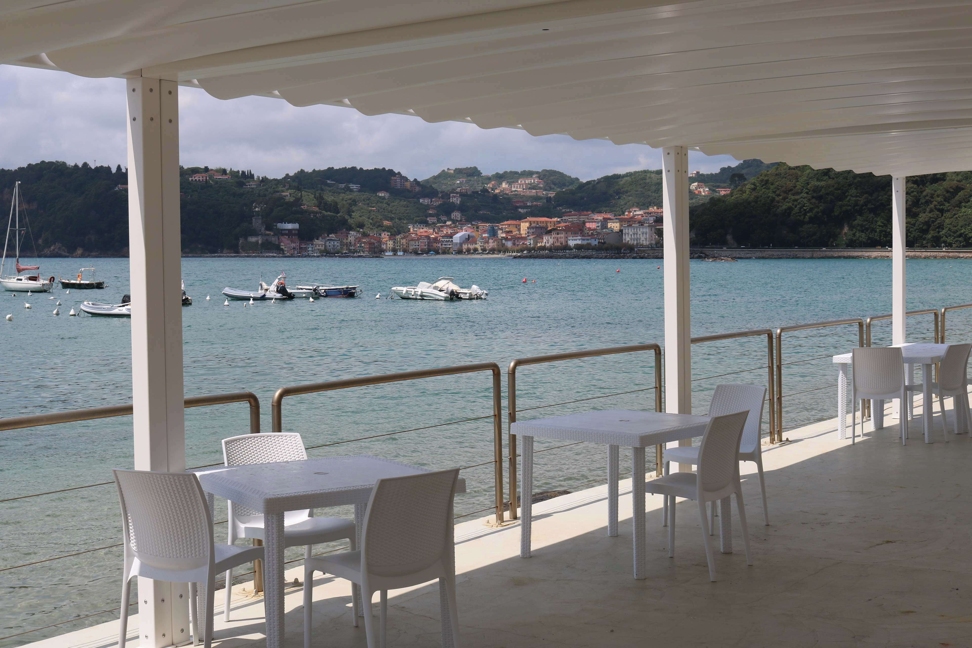 A covered outdoor seating area with tables and chairs overlooking a body of water with boats and a distant town.